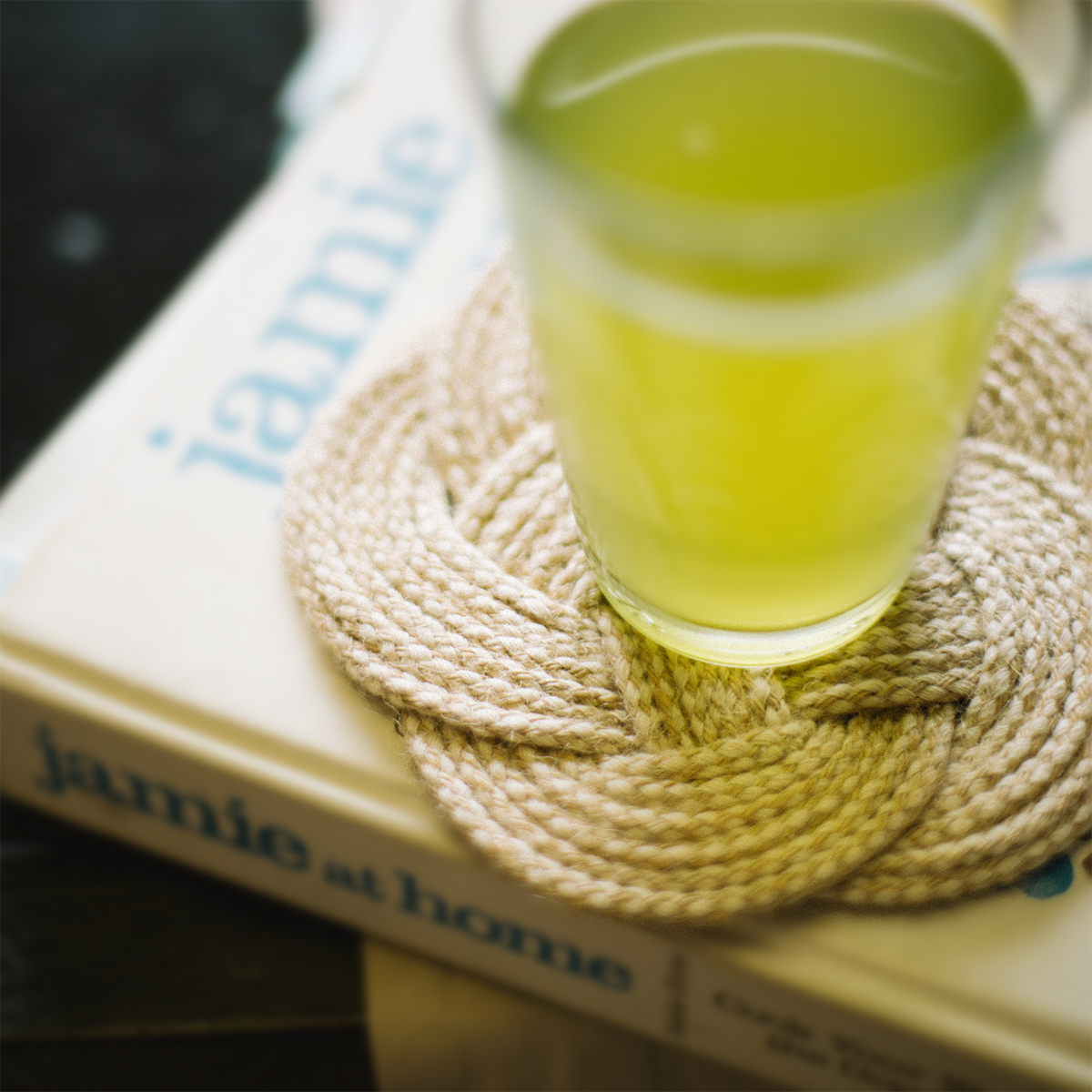Glass of yellow-green drink on woven coaster atop cookbook jamie at home