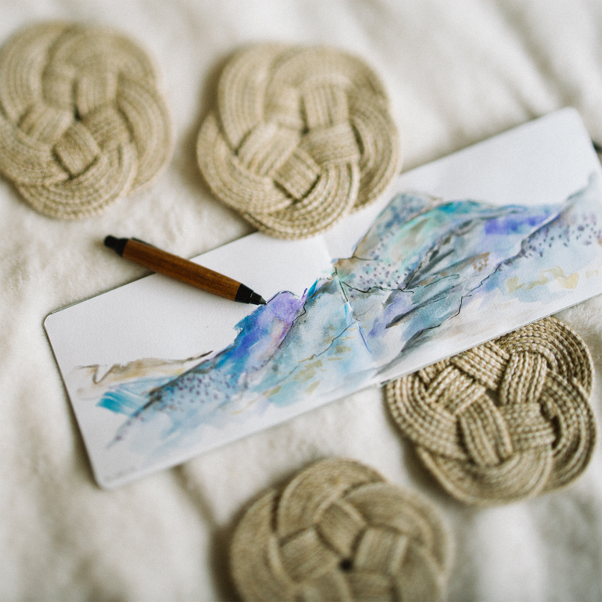 Watercolor sketch of mountains in a notebook with a wooden pen, surrounded by woven coasters.