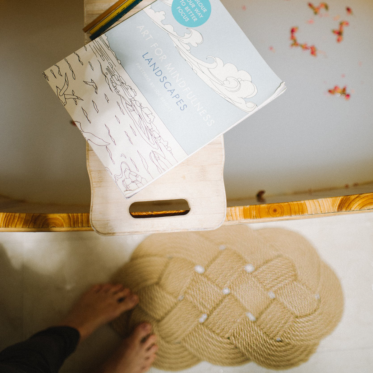 Book titled 'Art for Mindfulness Landscapes' on a wooden bath tray, feet on braided mat nearby