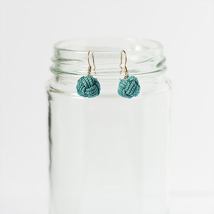 Pair of teal crochet earrings with gold hooks displayed on a clear glass jar, minimalist style.